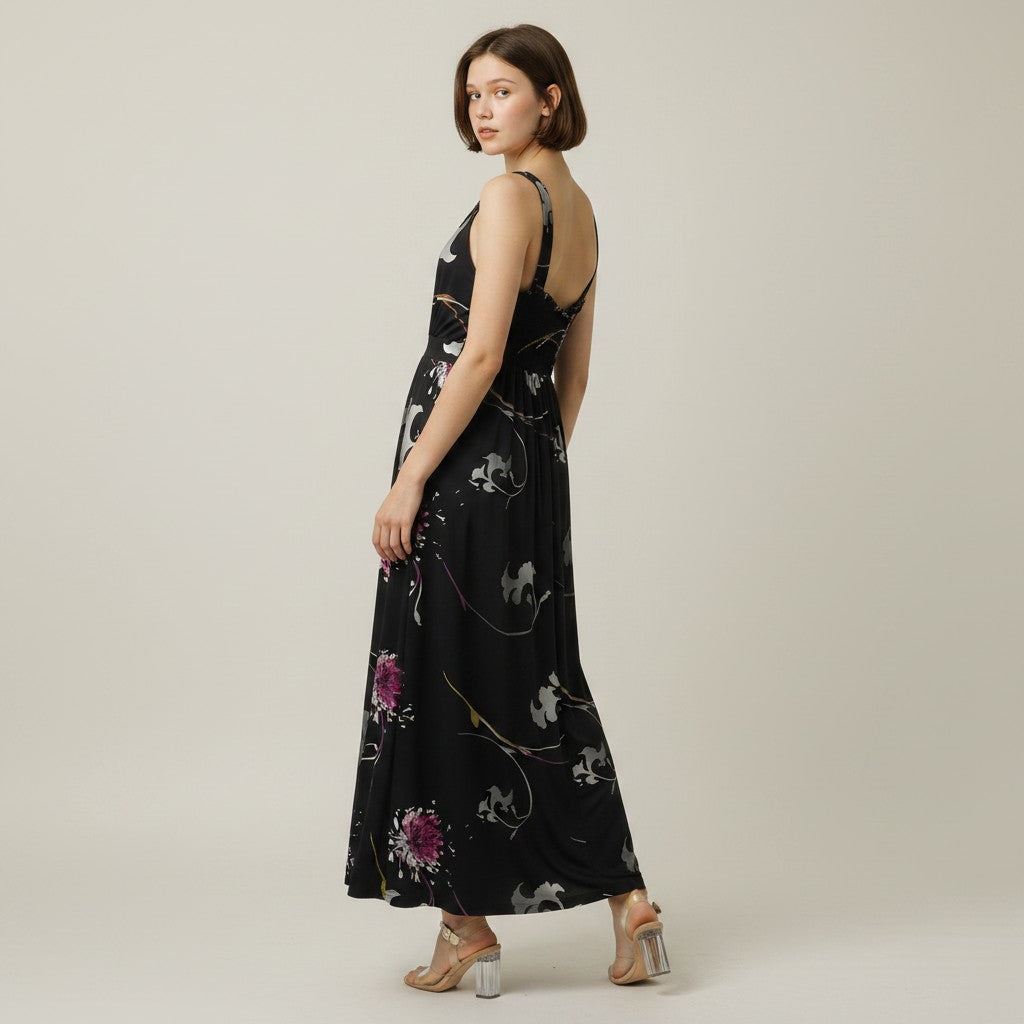 A woman modeling a sleeveless black maxi dress with a white floral and vine pattern, shown from a three-quarters back view against a neutral studio background.