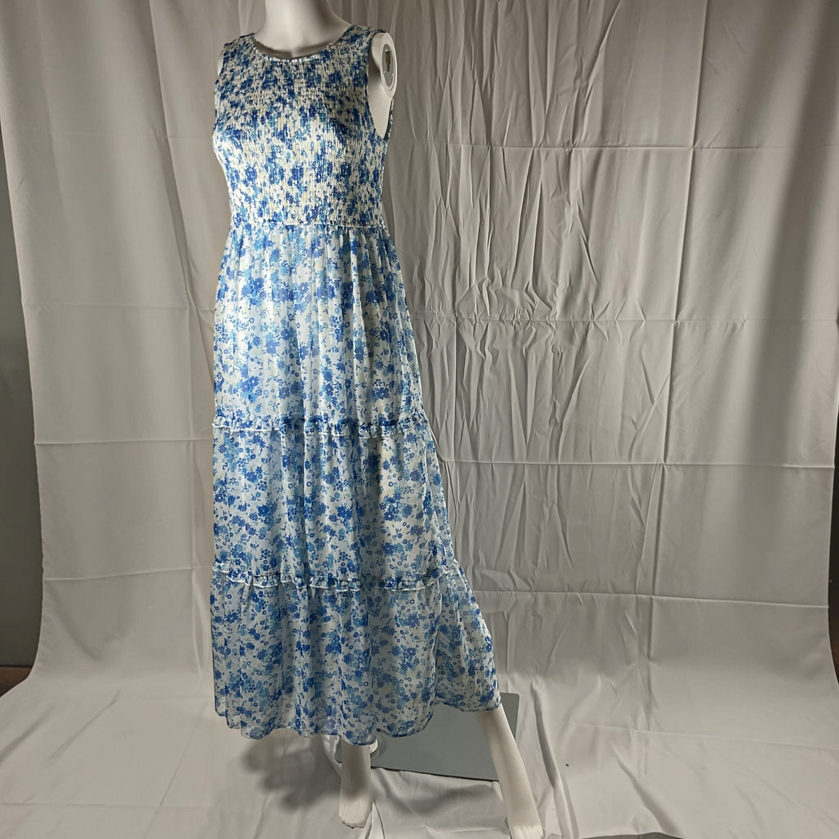 A sleeveless tiered maxi dress with a blue and white floral pattern, displayed on a white mannequin against a wrinkled white fabric backdrop.