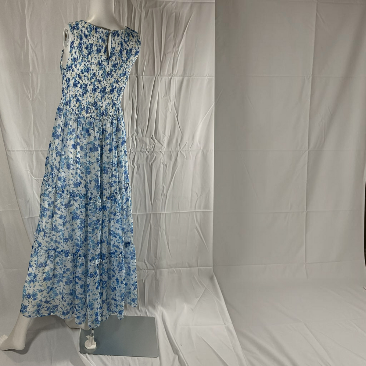 A sleeveless, tiered maxi dress with a blue and white floral pattern, displayed on a mannequin against a wrinkled white fabric backdrop.