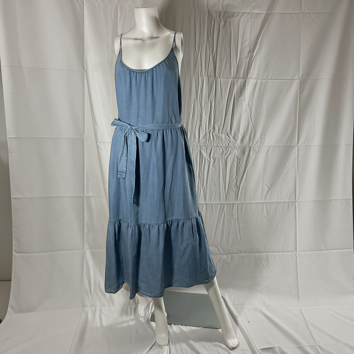 A blue sleeveless midi dress displayed on a white mannequin against a wrinkled white backdrop. The dress has a scoop neckline, thin shoulder straps, a matching fabric tie belt at the waist, and a tiered skirt.