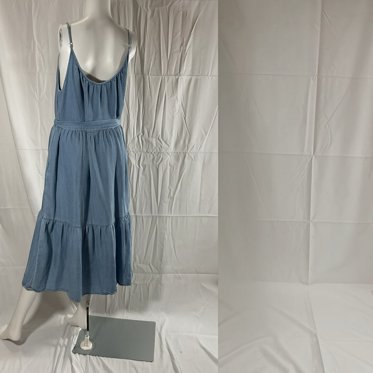A sleeveless tiered midi dress in dusty blue denim-like fabric displayed on a white mannequin against a wrinkled white studio backdrop.