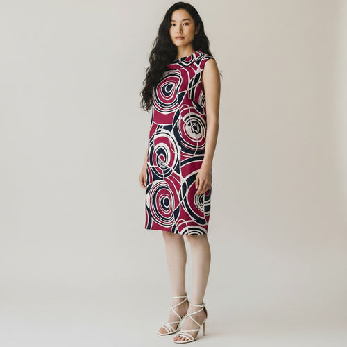 A woman modeling a sleeveless high-neck dress with a bold abstract spiral pattern in magenta, black, and white, paired with white strappy high-heeled sandals against a plain light beige studio background.