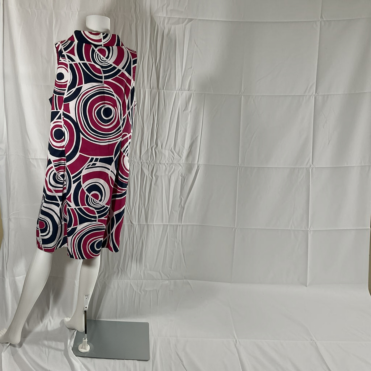 A sleeveless mock-neck dress with a bold abstract circular pattern in magenta, navy, black, and white, displayed on a white mannequin against a wrinkled white fabric backdrop.