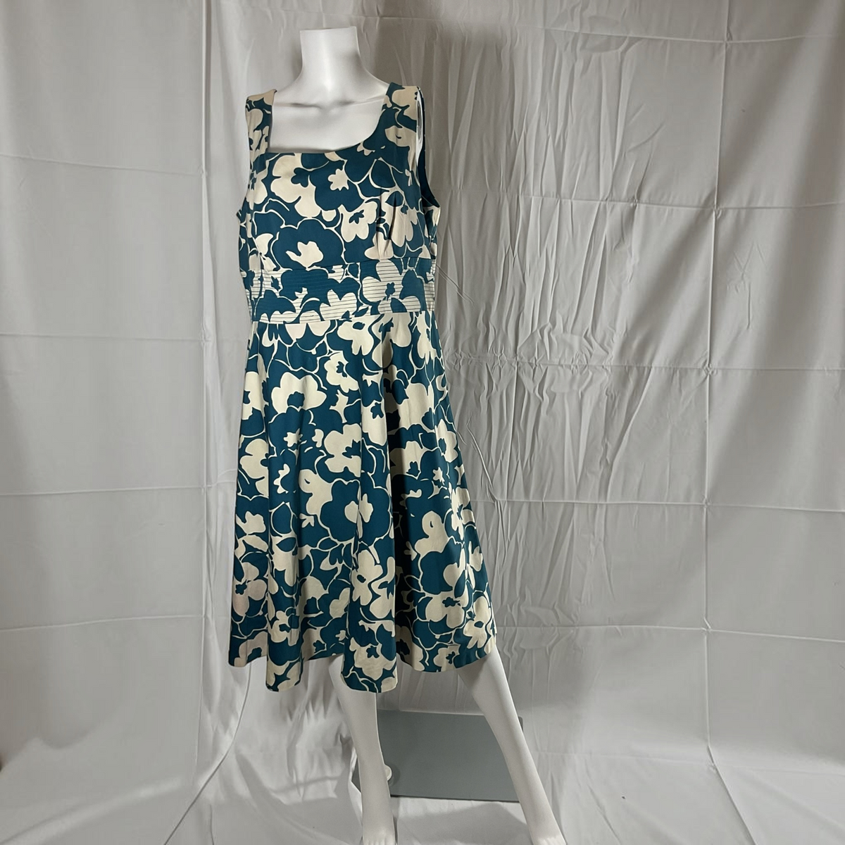 A sleeveless A-line midi dress with a blue and cream floral pattern, displayed on a white mannequin against a wrinkled white fabric backdrop.