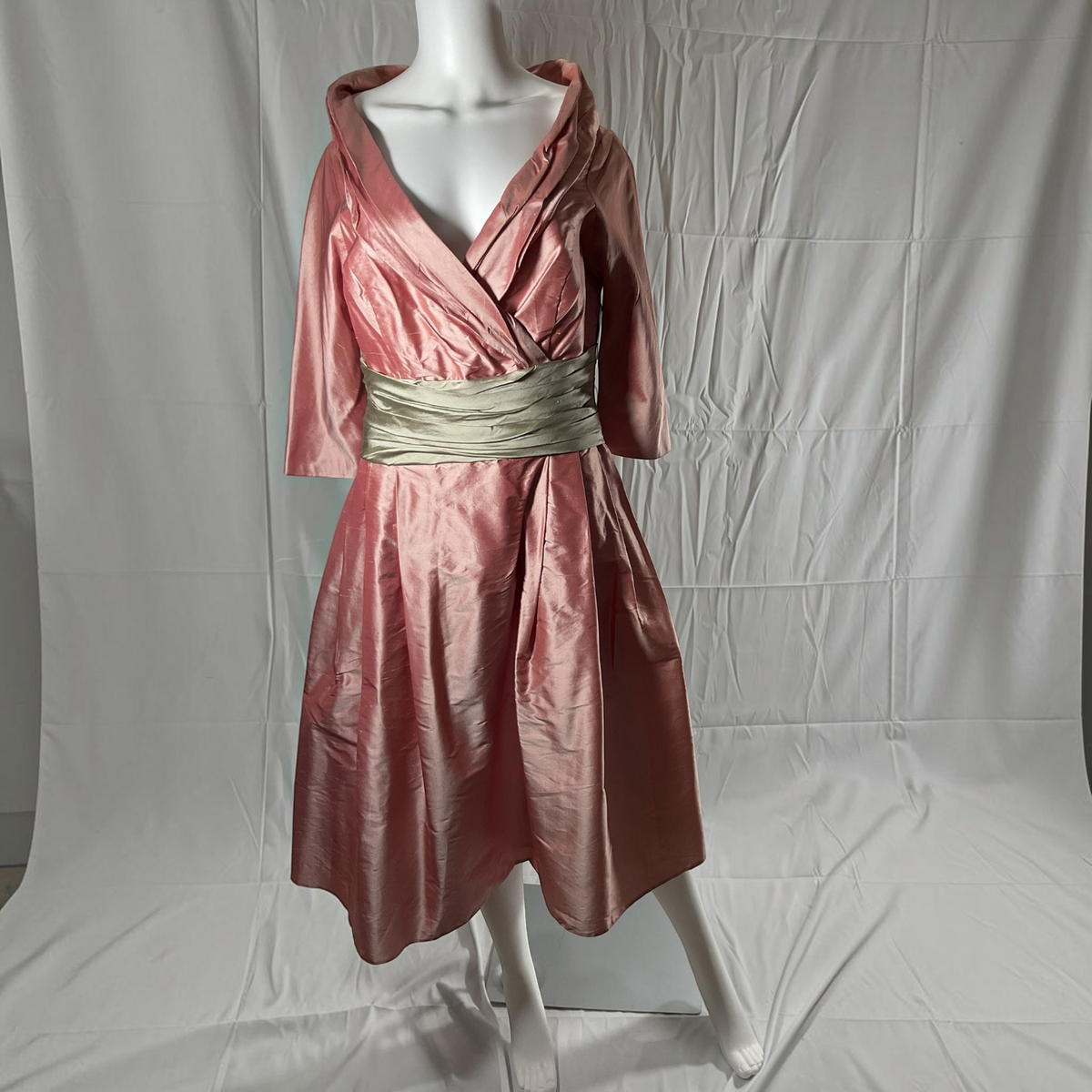 A pink satin-like hooded dress displayed on a white mannequin against a wrinkled white backdrop. The dress has three-quarter sleeves and a wide metallic gold sash at the waist.