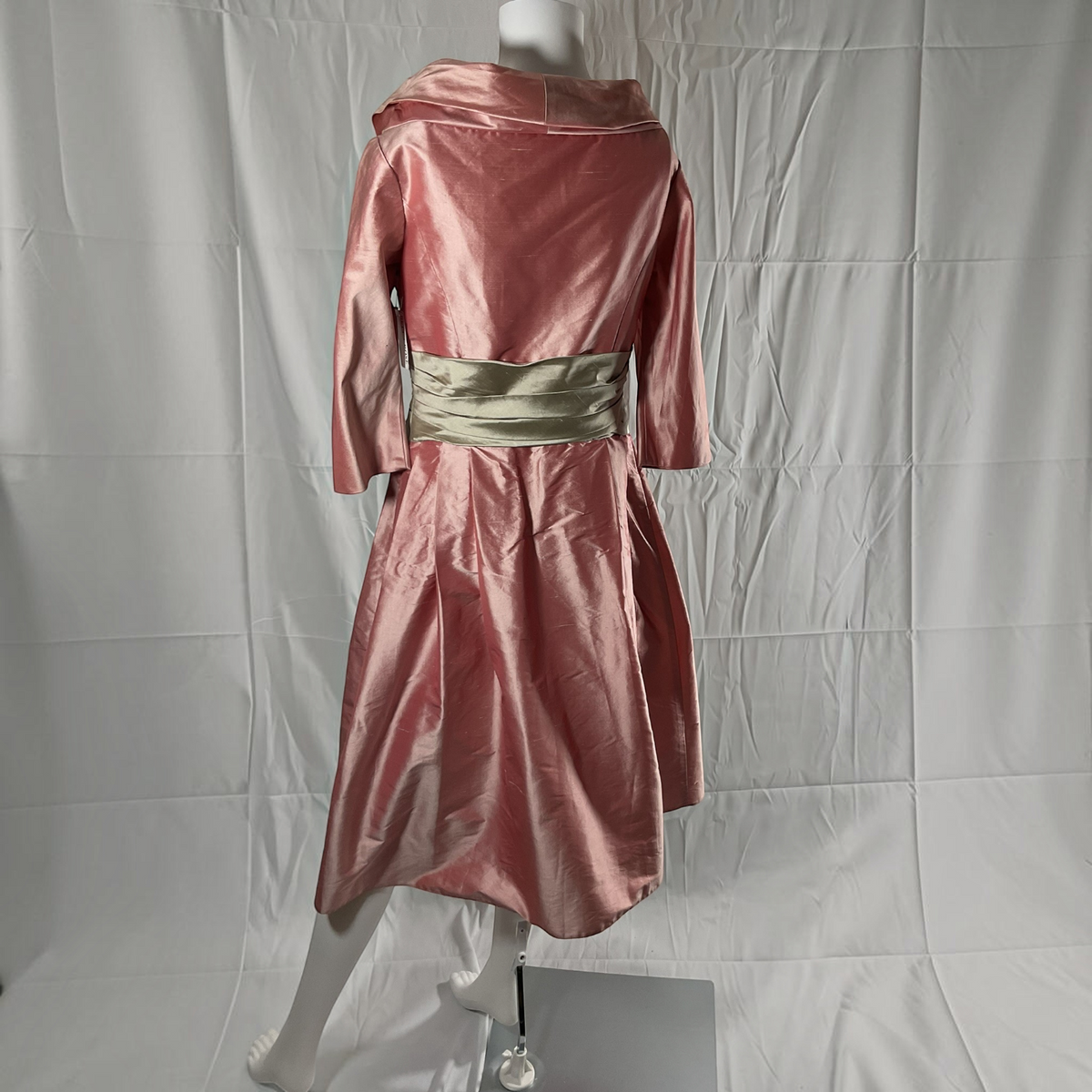 A pink satin-like hooded dress displayed on a white mannequin against a wrinkled white backdrop. The dress has three-quarter sleeves and a wide metallic gold sash at the waist.