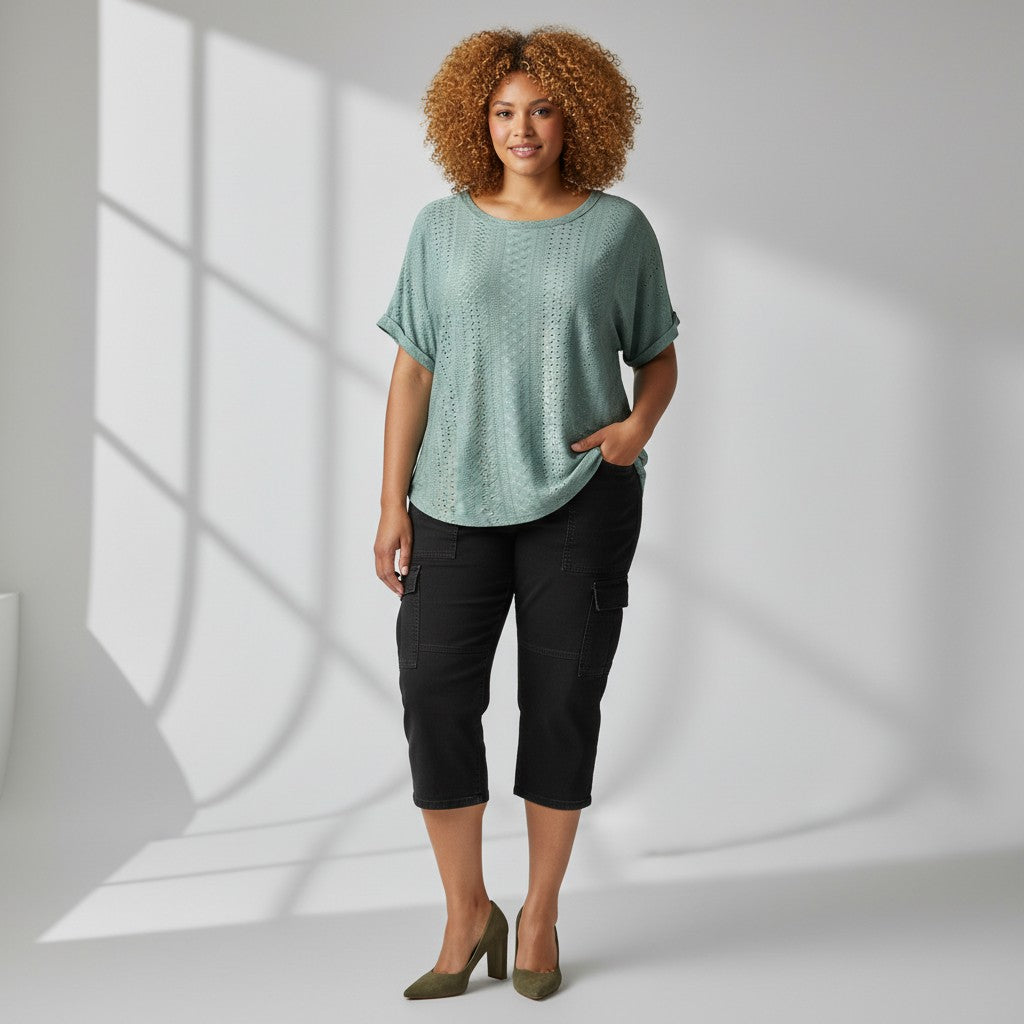 A woman modeling a sage green short-sleeve knit top with black cropped cargo pants and olive green heels, standing in a bright studio with window shadow patterns on a white background.