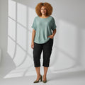 A woman modeling a sage green short-sleeve knit top with black cropped cargo pants and olive green heels, standing in a bright studio with window shadow patterns on a white background.