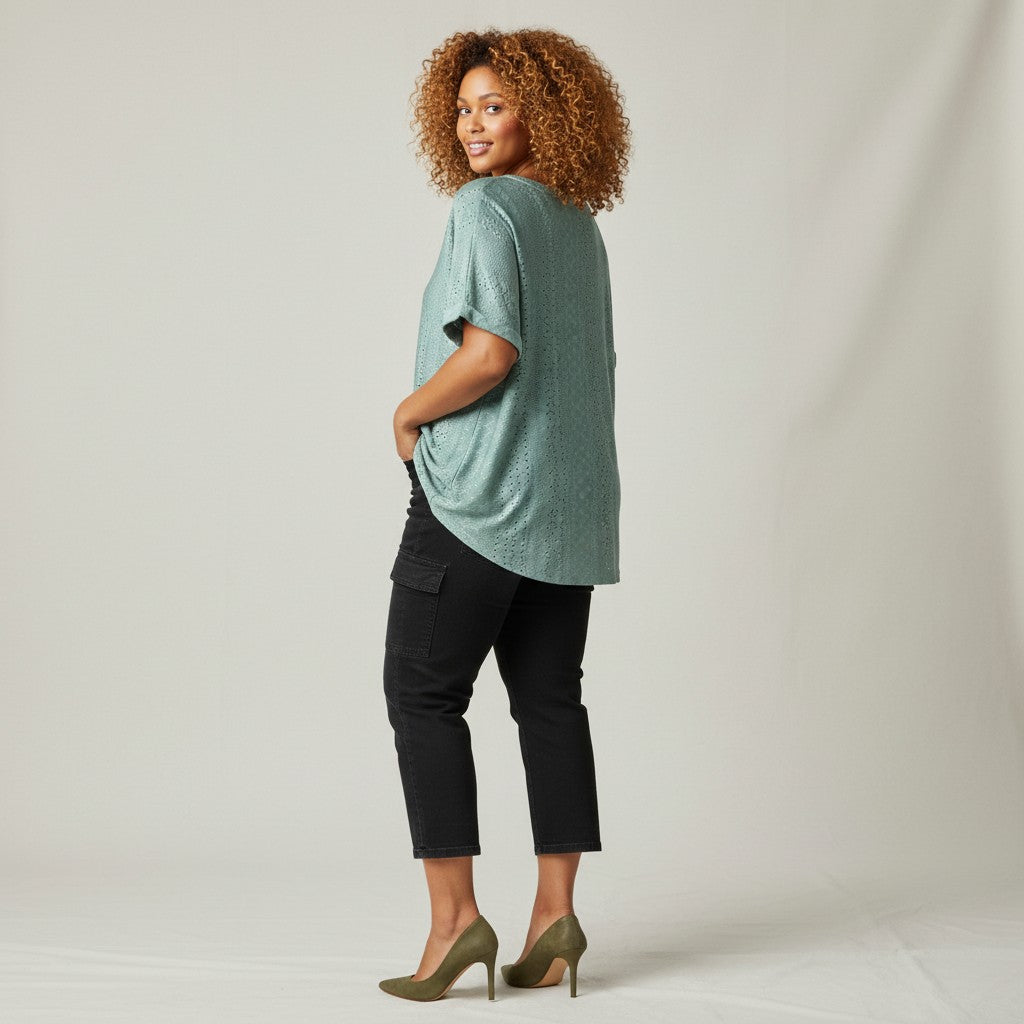 A woman with curly light brown hair is shown from the back wearing a sage green short-sleeve top, black cropped pants, and olive green heels against a light gray studio backdrop.