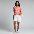 A woman modeling a pink and orange patterned long-sleeve top with white shorts and pink sneakers, standing against a light gray studio background.