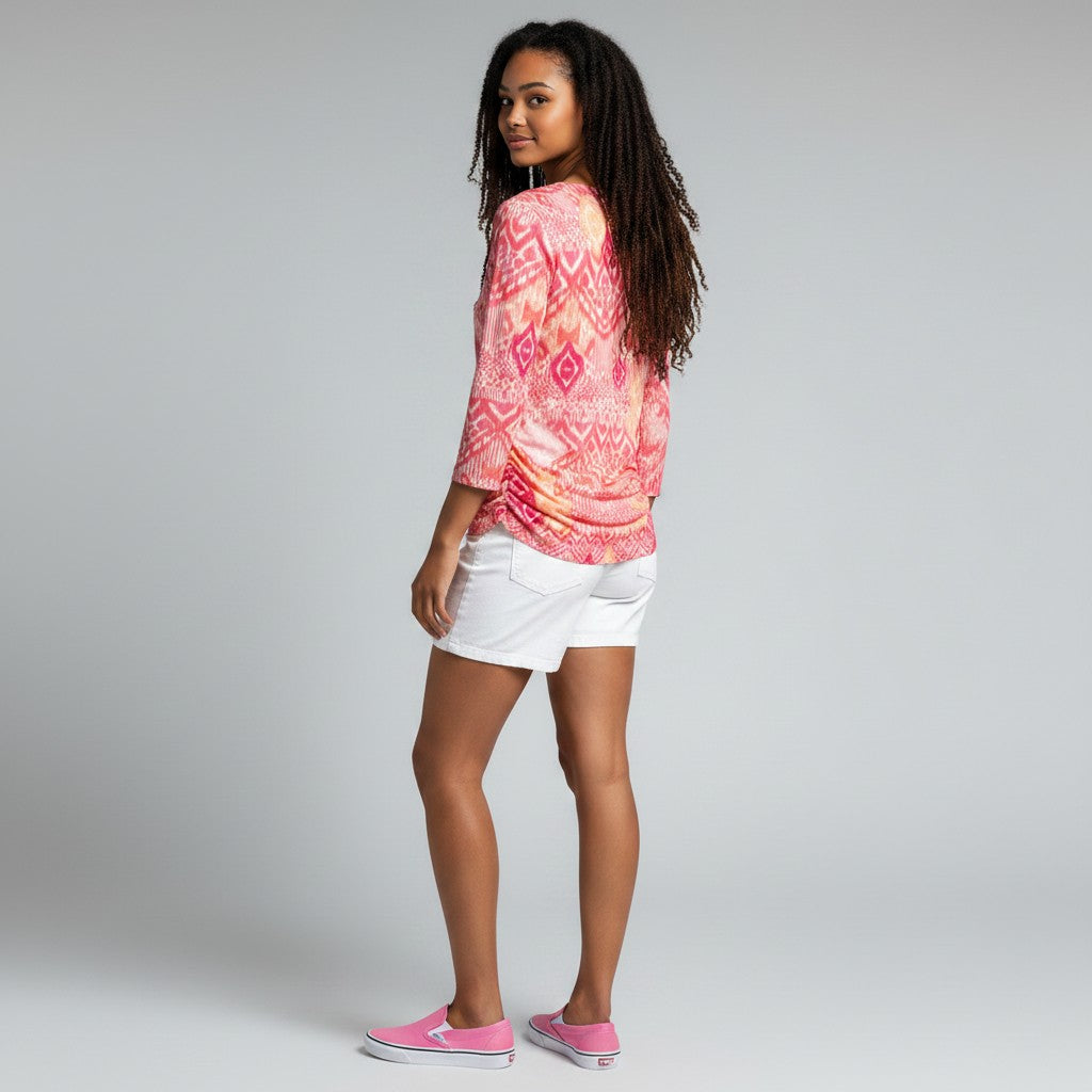 A woman stands in profile wearing a pink and coral patterned long-sleeve top, white shorts, and pink slip-on shoes against a light gray studio background.