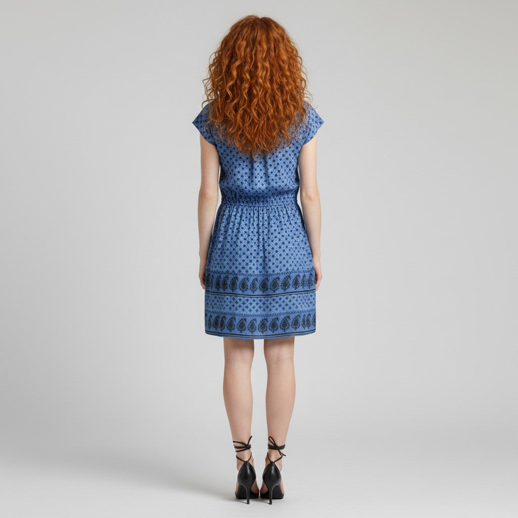 A woman with long curly red hair models a blue patterned sleeveless dress with a smocked bodice and a tiered skirt, paired with black strappy high-heeled sandals, against a light gray studio background.