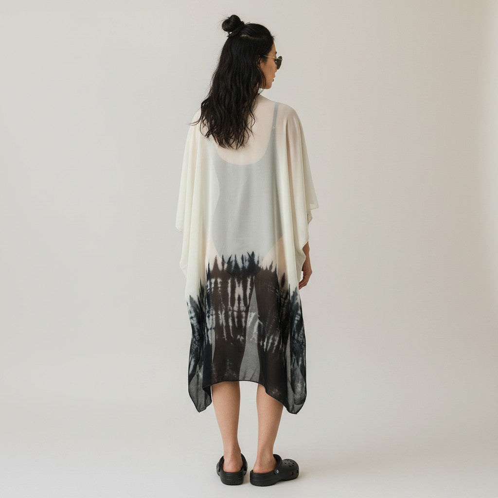 A woman viewed from behind wearing a loose, semi-sheer kaftan-style cover-up dress with a white and black tie-dye pattern, set against a plain light gray studio background.