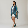 A woman models a lightweight draped shawl with an abstract watercolor pattern in shades of teal, blue, and green, worn over a white dress against a neutral studio background.