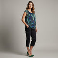 A woman modeling a sleeveless scoop-neck top with a blue and green geometric pattern, paired with black cropped pants and green heels, against a solid gray studio background.