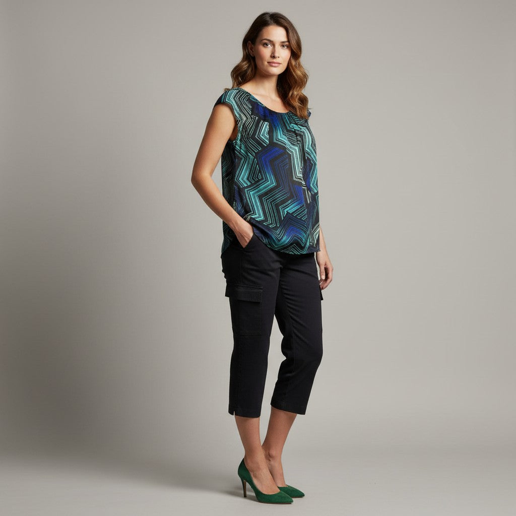 A woman modeling a sleeveless scoop-neck top with a blue and green geometric pattern, paired with black cropped pants and green heels, against a solid gray studio background.