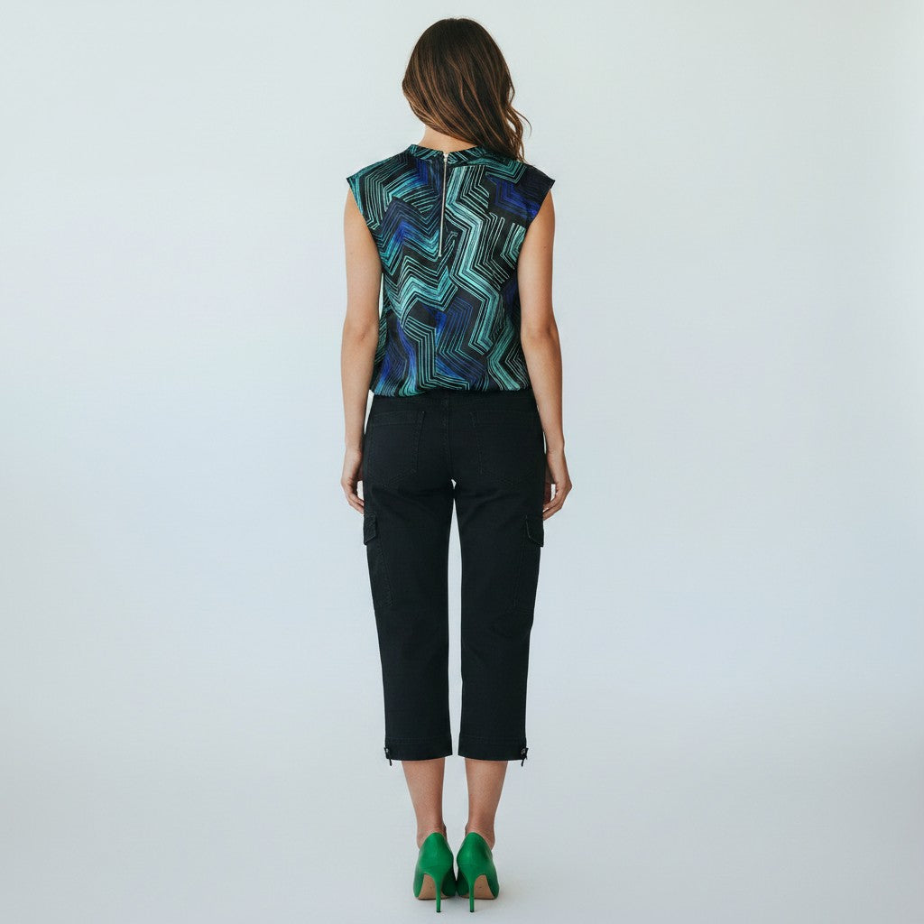 A woman viewed from the back wearing a sleeveless top with a blue, green, and black geometric zigzag pattern, paired with black cropped pants and green heels, against a plain light gray studio background.