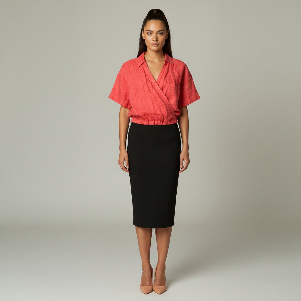 A woman stands against a light gray studio background wearing a coral pink wrap-style short-sleeve blouse and a black knee-length pencil skirt with thin white side stripes, paired with nude heels.