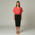 A woman stands against a light gray studio background wearing a coral pink wrap-style short-sleeve blouse and a black knee-length pencil skirt with thin white side stripes, paired with nude heels.