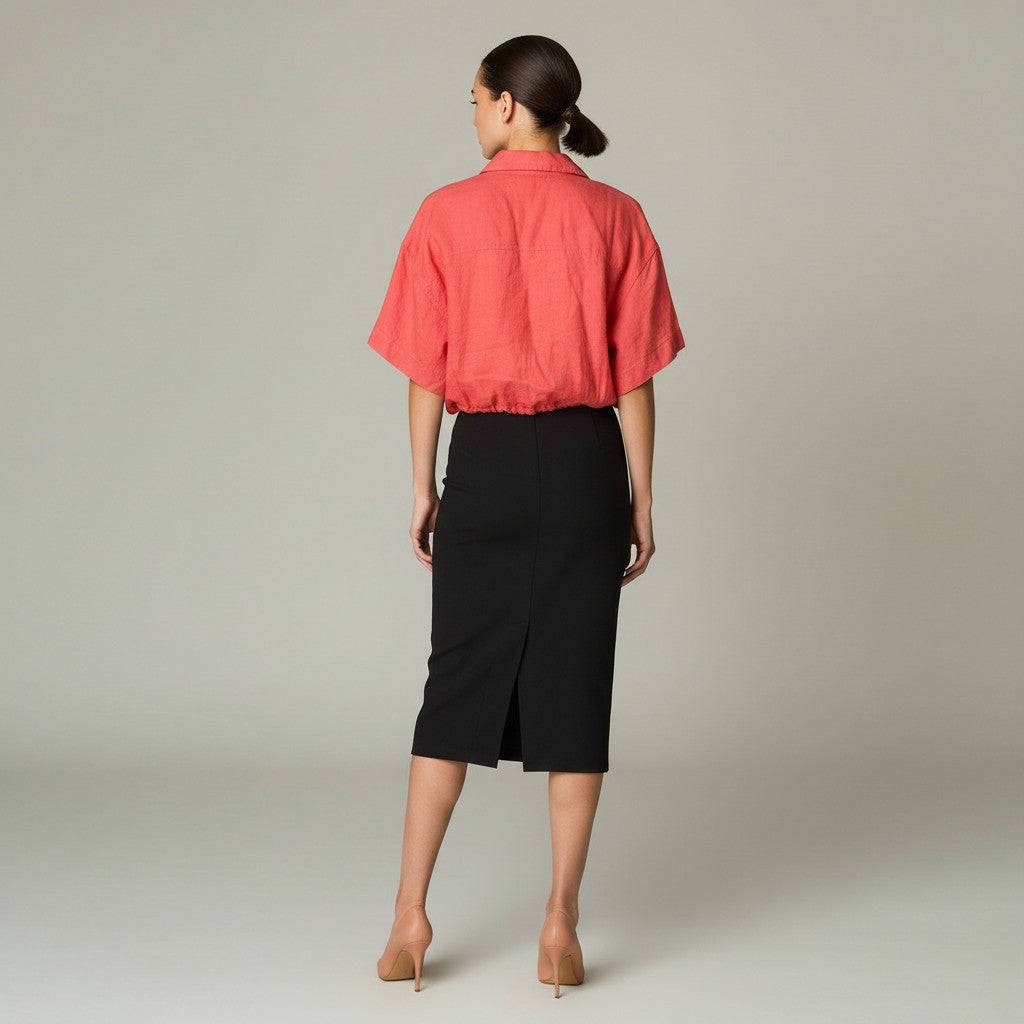 A woman is shown from the back wearing a coral pink short-sleeve button-up shirt and a black knee-length skirt, standing against a neutral light gray studio background.