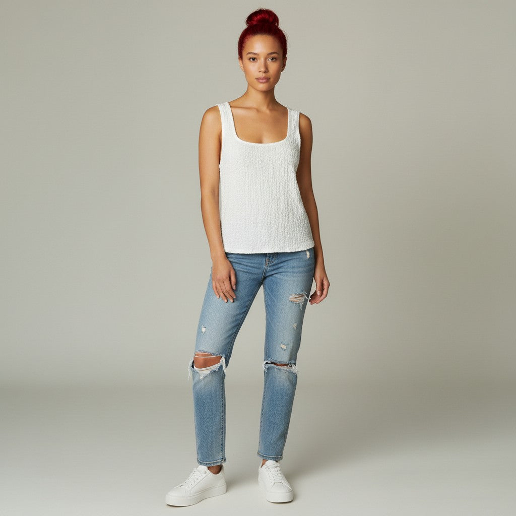 A woman modeling a white ribbed knit tank top with light-wash distressed blue jeans and white sneakers, standing against a neutral light gray studio background.