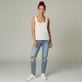 A woman modeling a white ribbed knit tank top with light-wash distressed blue jeans and white sneakers, standing against a neutral light gray studio background.
