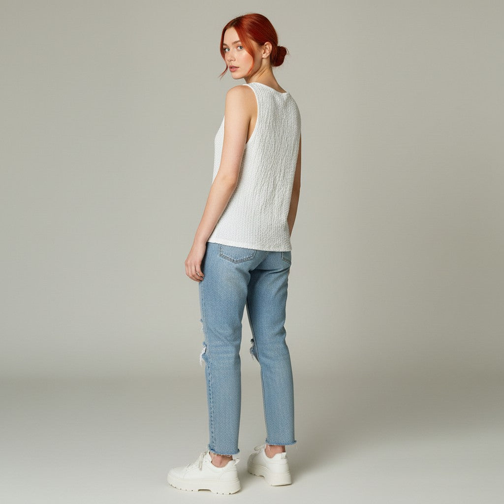 A woman with red hair tied back is shown from the back wearing a white sleeveless textured knit top, light blue jeans, and white sneakers against a neutral light gray studio background.