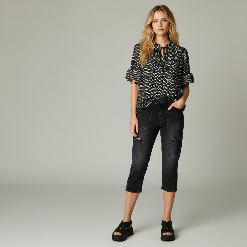 A woman modeling a black and white patterned short-sleeve blouse with a tie-neck detail, paired with black cropped jeans and black platform sandals, against a neutral gray studio background.