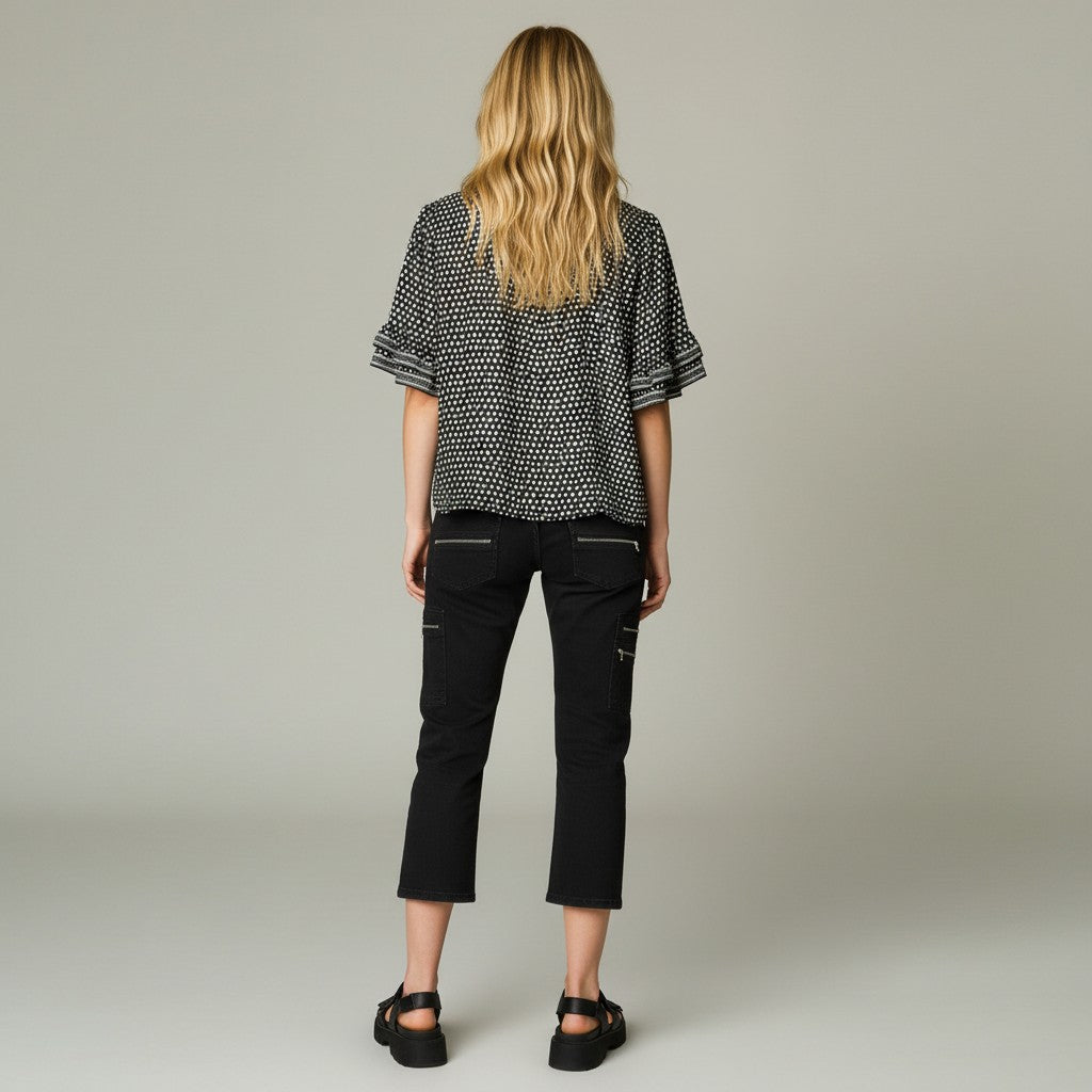 A woman viewed from behind wearing a black and white patterned short-sleeve blouse, black cropped pants, and black sandals, standing against a plain light gray studio background.