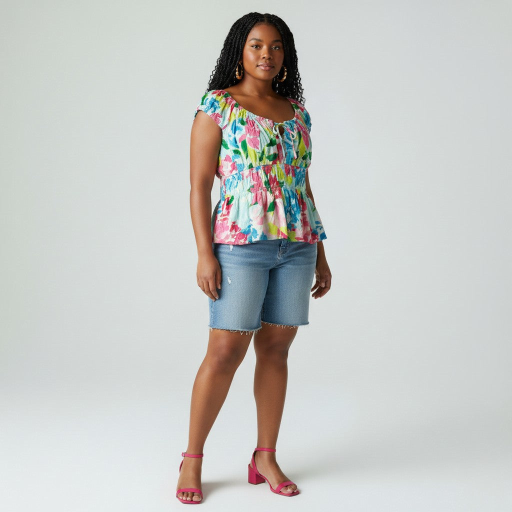 A woman modeling a colorful floral print peplum top with denim shorts and pink heels, standing against a light gray studio background.