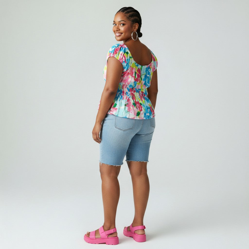 A woman modeling a colorful, short-sleeved scoop-neck top with a vibrant watercolor-like print in shades of blue, pink, yellow, and green. She is also wearing light-wash denim shorts and bright pink platform sandals, photographed against a solid light gray studio background.
