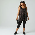 A woman modeling a sleeveless black and dark gray patterned tank top, paired with black capri leggings, black sneakers, and a black baseball cap, holding a black water bottle against a light gray studio background.