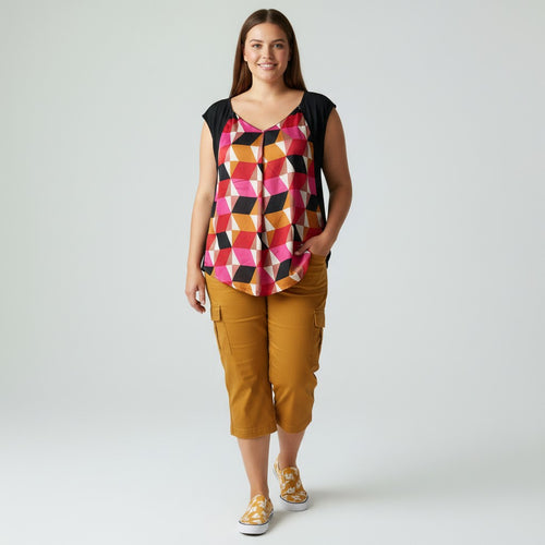 A woman modeling a sleeveless top with a colorful geometric diamond pattern in pink, orange, yellow, and black over a black underlayer, paired with mustard cargo capri pants and yellow patterned sneakers, against a light gray studio background.