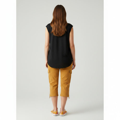 A woman viewed from behind wearing a black sleeveless top and mustard yellow cropped cargo pants, standing against a light gray studio background.