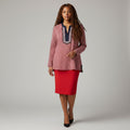 A woman modeling a textured dusty pink long-sleeve tunic top with a navy and silver embroidered V-neckline, paired with a red pencil skirt and black heels, against a neutral gray studio background.