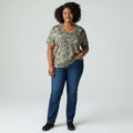 A woman modeling a short-sleeve V-neck t-shirt with a green and pink floral pattern, paired with dark blue jeans and black shoes, standing against a light gray studio background.