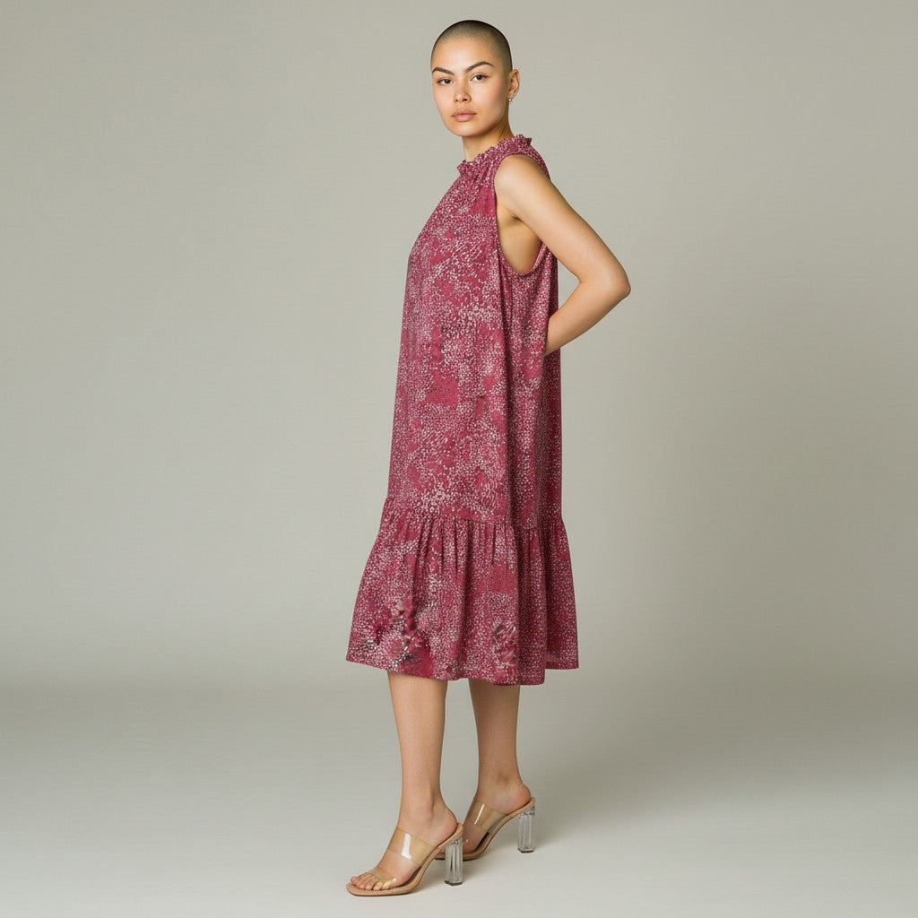 A woman modeling a sleeveless tiered midi dress with a pink and white floral pattern, paired with clear high-heeled sandals, against a neutral gray studio background.