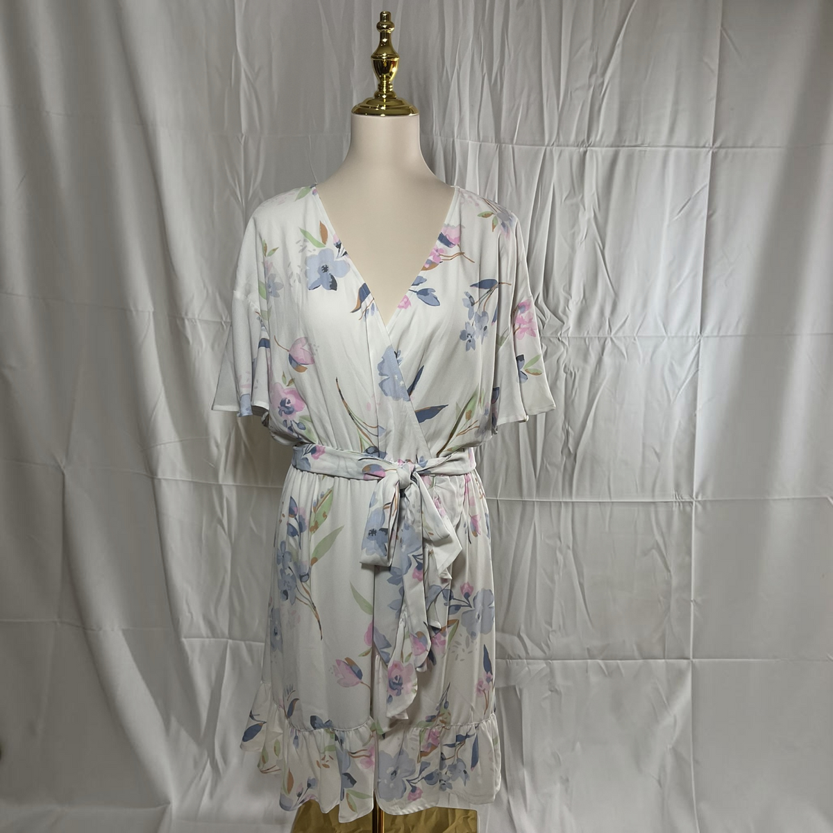 A short-sleeve wrap dress with a tie waist, featuring a watercolor floral print in shades of blue, pink, and green on a white base, displayed on a mannequin against a wrinkled white fabric backdrop.