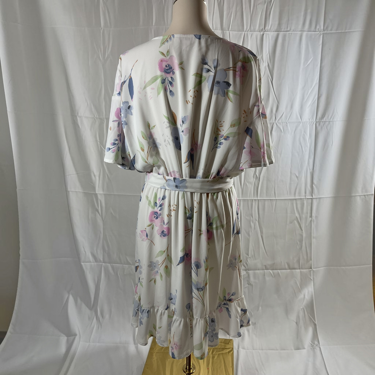 A short-sleeve floral dress displayed on a mannequin against a white fabric backdrop. The dress has a cream base with a watercolor-style print in pink, blue, and green, and features a matching fabric tie belt at the waist.