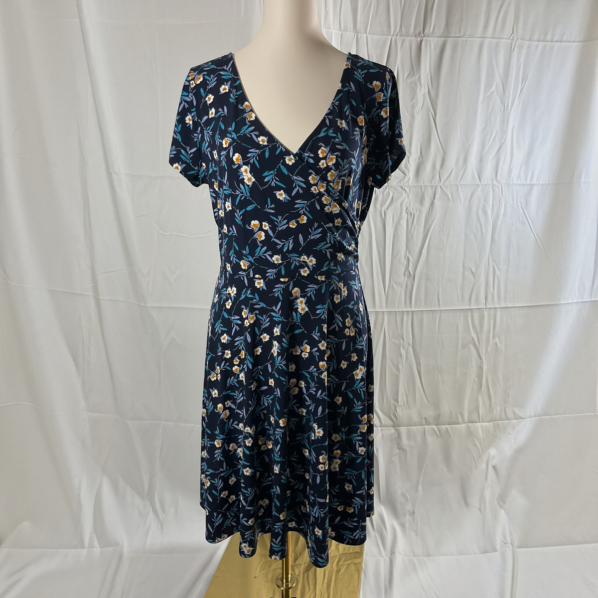 A navy blue short-sleeve V-neck dress with a white and yellow floral print, displayed on a mannequin against a white wrinkled fabric backdrop.