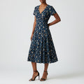 A woman models a navy blue midi dress with a white and beige floral pattern, featuring short sleeves, a V-neckline, and a flared skirt, paired with black strappy high-heeled sandals against a light gray studio background.