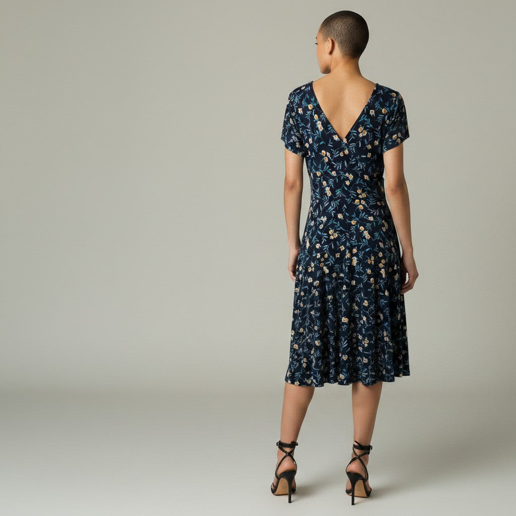 A woman viewed from the back wearing a navy wrap-style midi dress with a beige and white floral pattern, paired with black strappy heels, against a neutral gray studio background.