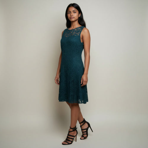 A woman modeling a sleeveless, knee-length dark teal lace dress with a scoop neckline and black strappy high-heeled sandals, standing against a plain light beige studio background.