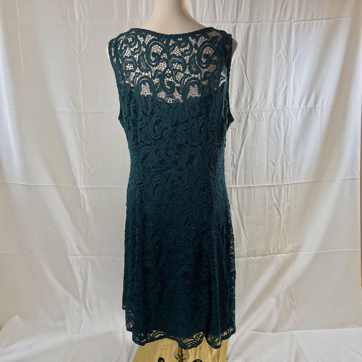 A sleeveless dark teal lace dress displayed on a mannequin torso against a white fabric backdrop.