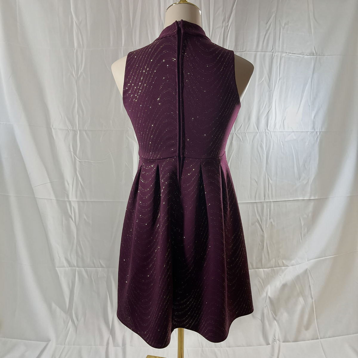 A sleeveless high-neck A-line dress in deep plum purple with a subtle wavy pattern and small silver reflective dots, displayed on a dress form against a wrinkled white fabric backdrop.