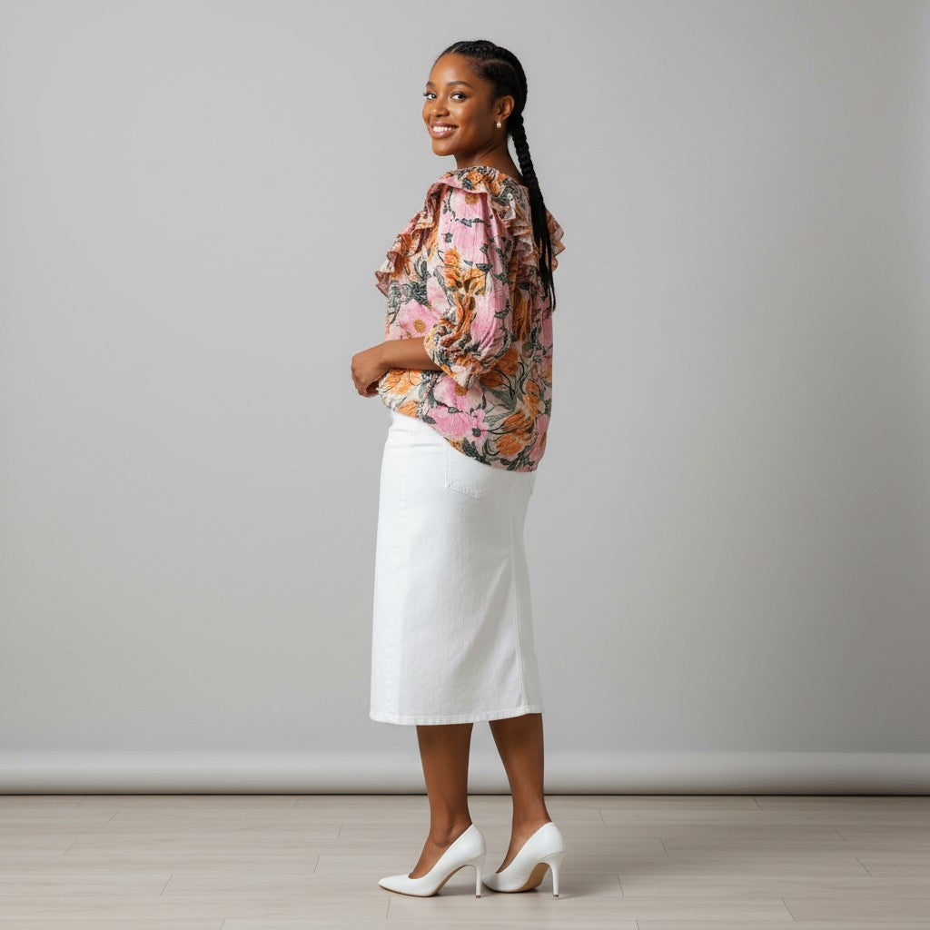 A woman stands in profile wearing a pink floral blouse with ruffled details and a white midi skirt, paired with white heels, against a plain light gray studio background.