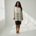A woman stands in a bright white studio wearing a cream lace tunic top with a black skirt and white heels. The background features soft window shadow patterns cast across the floor and wall.