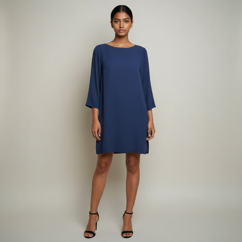 A woman modeling a navy blue shift dress with three-quarter sleeves and a boat neckline, paired with black strappy high-heeled sandals, against a neutral light gray studio background.
