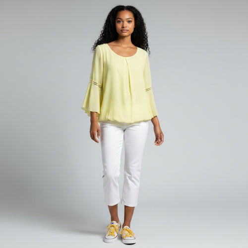 A woman modeling a pale yellow three-quarter sleeve blouse with decorative gold embroidery on the cuffs, paired with white cropped pants and yellow sneakers, against a light gray studio background.