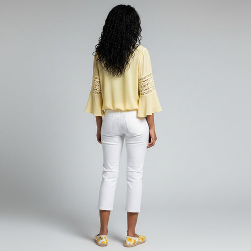 A woman shown from the back wearing a pale yellow blouse with lace sleeve details and white cropped pants, standing against a light gray studio background.